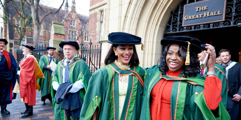 Features two bright and smiling new graduates in green and red robes outside of the Great Hall at the University of Leeds