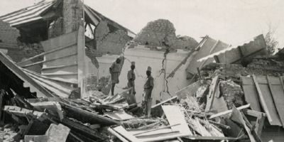 A sepia photo from 1935. Three men stand atop a pile of wood and brick rubble, in front of the single remaining wall of a house.