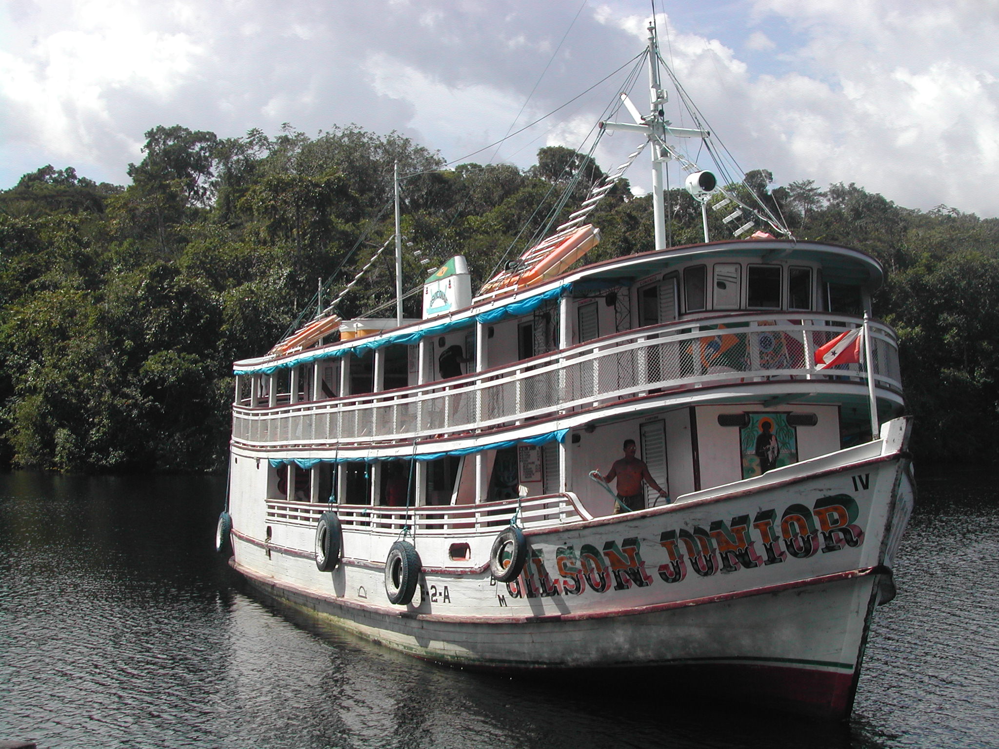 A large boat on the amazon river, with trees behind. Its name is painted on the side in bright colours, "Gilson Junior," and there are two people on the deck.