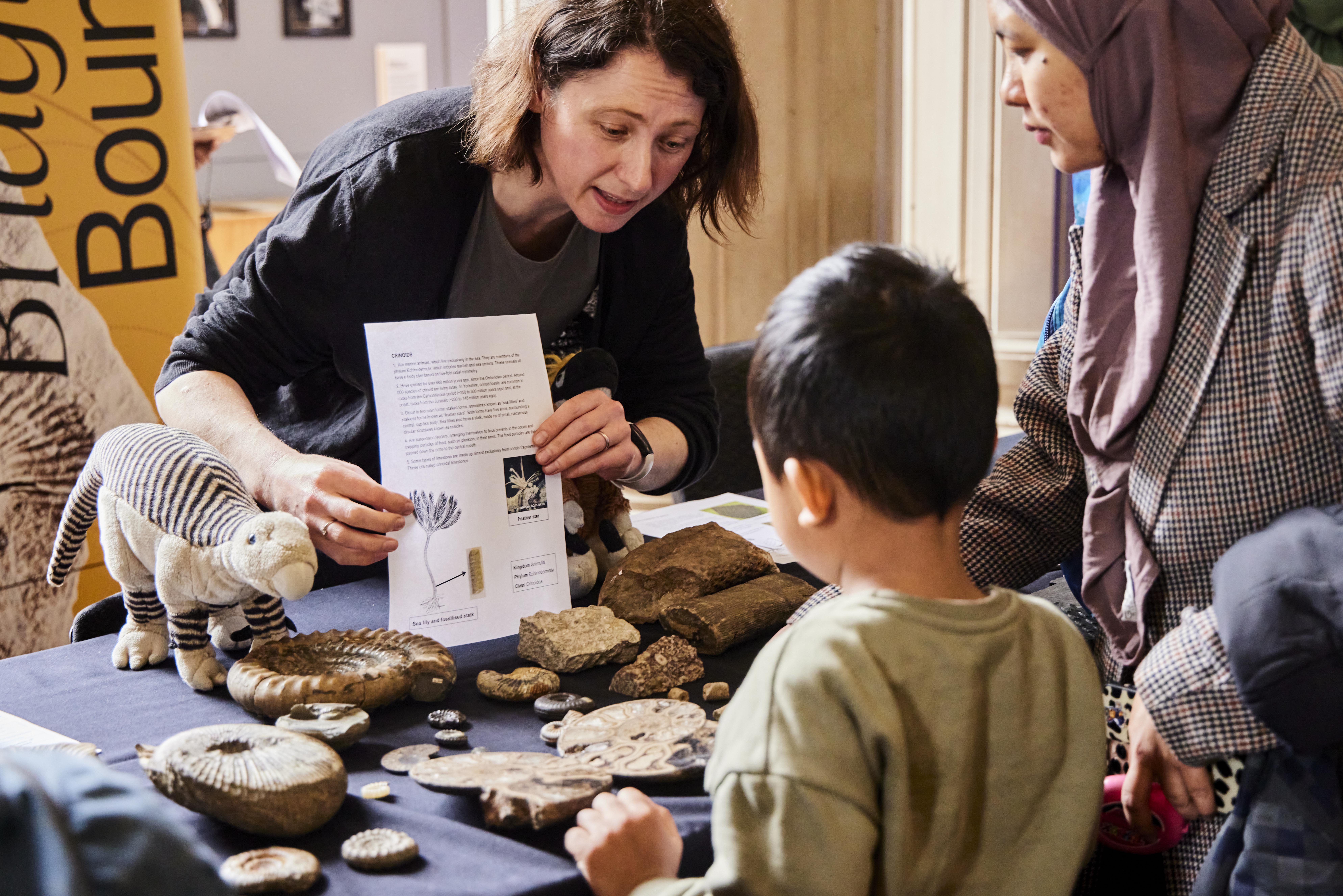 A researcher holds up a sheet of information about fossils to a child and parent. The table has fossils and a toy dinosaur on it.