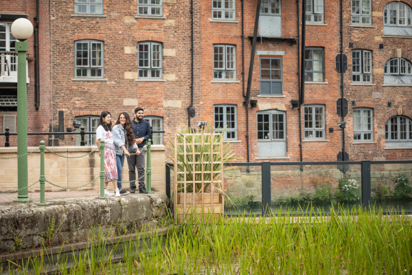 Three people stood looking into the River Aire with converted flats behind them.