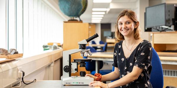 A female student sits beside a microscope.