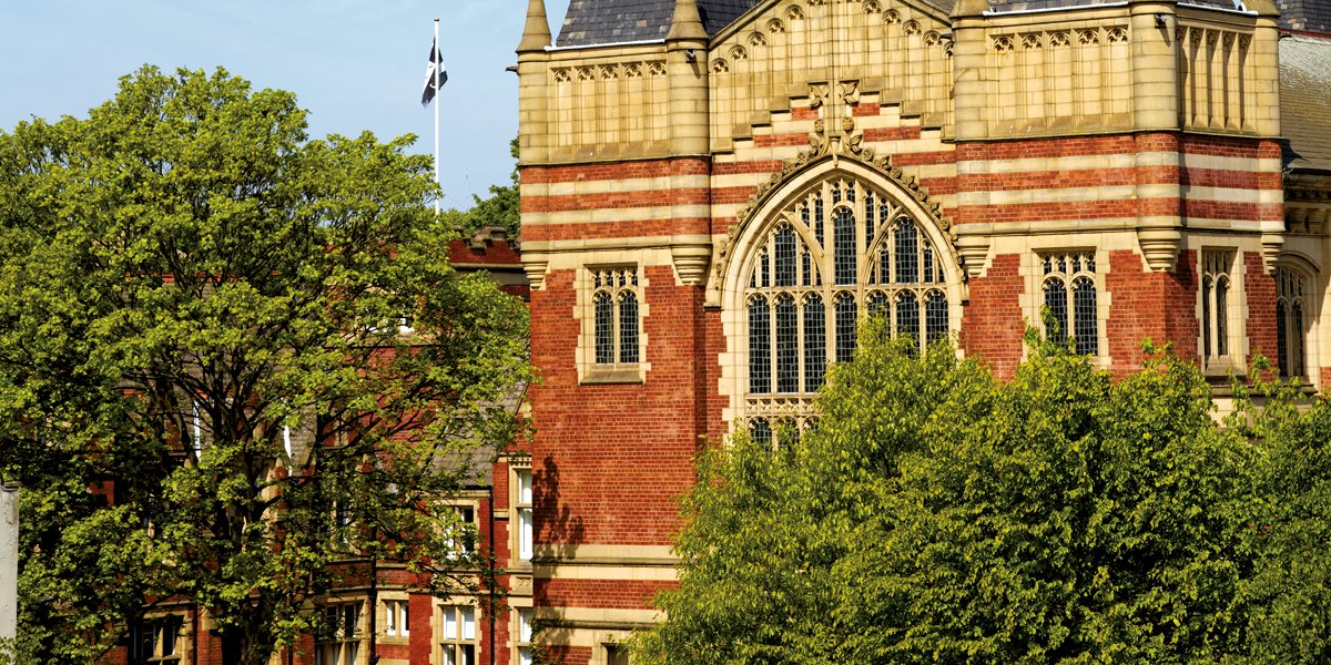 A cropped photograph of The Great Hall bathed in warm sunlight surround by leafy green trees, at the University of Leeds.
