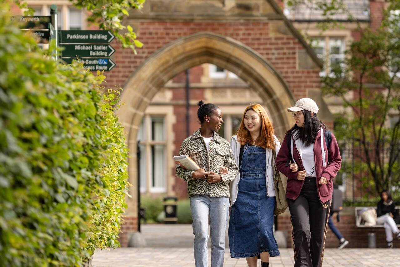 Three students walk and smile through a leafy university campus, under an archway. They seem engaged and happy, with books in hand.