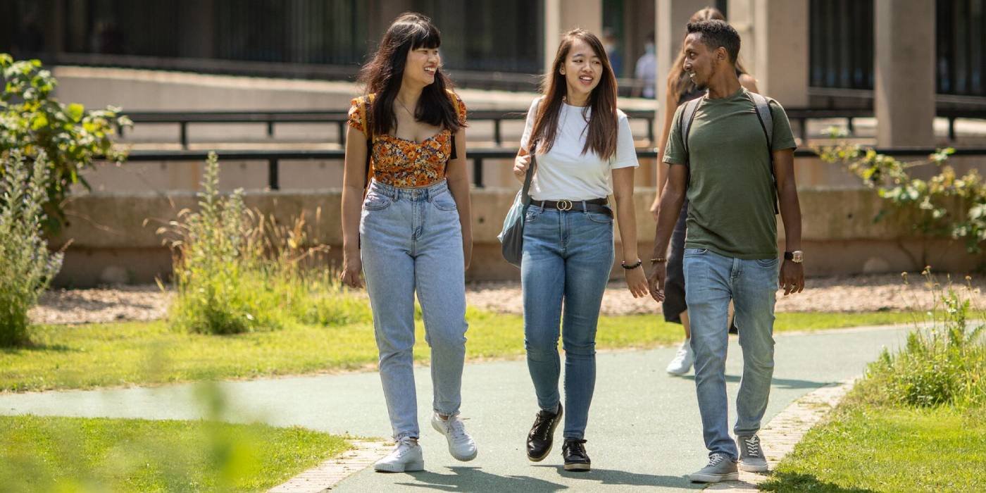 Three students walking through the Sustainable Garden on a sunny day at the University of Leeds. The students are walking along a green tarmac path, either side is green grass and plants.