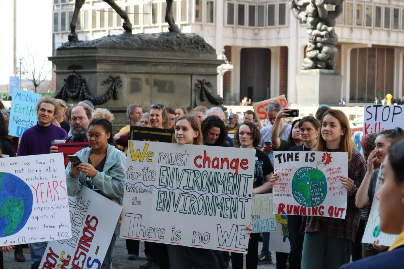 A crowd of people holding climate protest signs. One reads "We must change for the environment, without the environment there is no we." another says "time is running out".