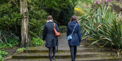 Two people walking on a path in a park, with trees and shrubs on either side