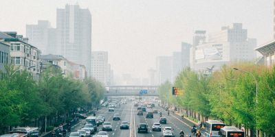 A road leading into Beijing, lined with trees and full with cars and buses. The city in the distance is foggy.