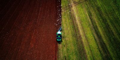 A birds-eye view of a field being ploughed by a tractor, with the left of the field showing brown tilled earth and the right side green. Seagulls fly behind the tractor.