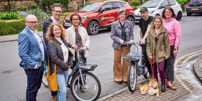 A group of people on a pavement, one holding a push bike, a dog sitting at another's feet. In the background are cars with 'Enterprise Car Club' branding.