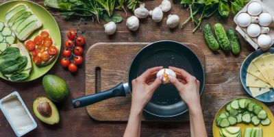 Two hands cracking an egg in a pan. Chopped vegetables and  mushrooms are on the table around it.