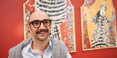 Artist Mohammad Barrangi smiles in front of works from 'One Night, One Dream, Life In the Lighthouse' displayed on a red wall