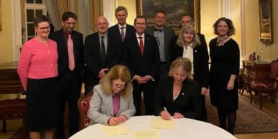 Two university leaders sit on a table signing a paper. Nine more stand behind them.