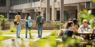 Three students stood with their back to the camera, in the Sustainable Garden on campus. It is a bright summers day. There are more students sat on a picnic table to the right foreground and greenery out of focus in the main foreground.