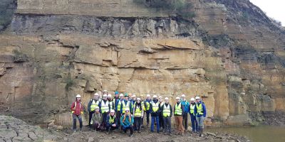 A group of researchers in hard hats and high vis jackets in a quarry, a wall of rock behind them