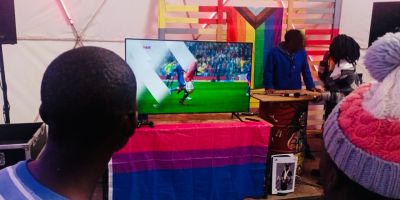 Someone playing Fifa seen from behind. A bisexual pride flag is under the screen and a progress pride flag is behind it.