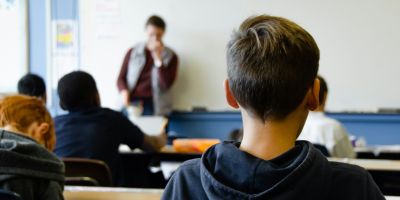 The back of a child's head as he sits in a classroom facing the teacher who is stood in front of a whiteboard.