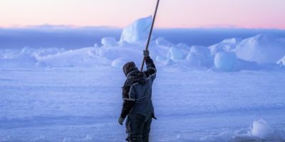 The icy landscape of Greenland with a low sun. Someone stands on the snow, holding a stick high in the air as if to put it through the ice.