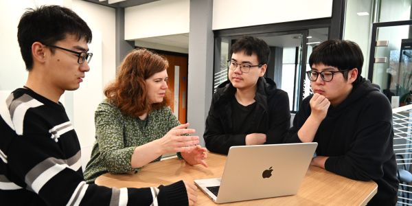 Three researchers sitting at a table with their supervisor, looking at a laptop together and talking about the research paper on the screen