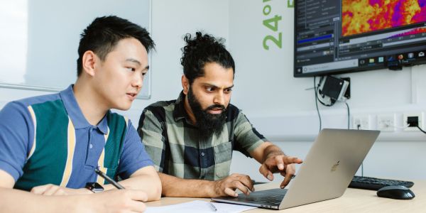 Two students sat a table with a laptop, one gesturing to the screen. Behind them there is the corner of a digital screen visible showing what appears to be a heat map.