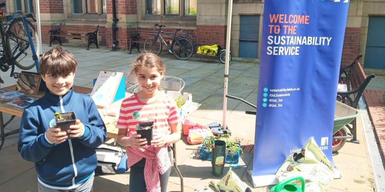 Two children smiling and holding plants in front of a Sustainability Service banner