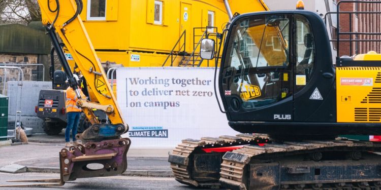 An industrial site with a yellow and black digger in front. A banner on the metal barrier says "Working together to deliver net zero on campus. Delivering net zero by 2030: CLIMATE PLAN".