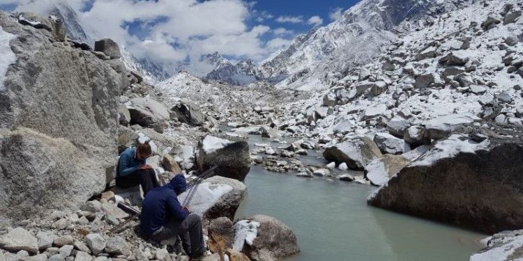 Two researchers sitting on rocks beside a glacier, icy mountains in the distance.