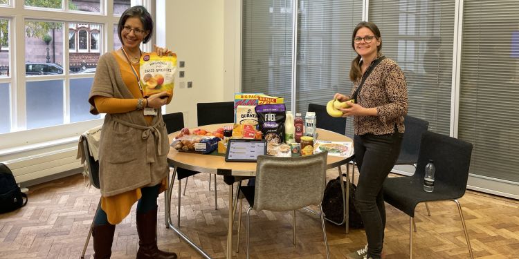 Yael Benn and Sally Moore at ESRC festival, standing in front of a table of food products and holding fruit.