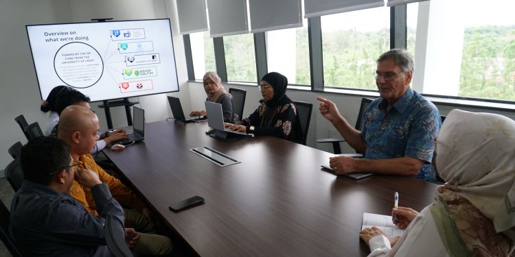 A bright meeting room with researchers from Indonesia and the UK sitting around a table and talking. A screen at the front has a presentation on that is titled "Overview on what we're doing"