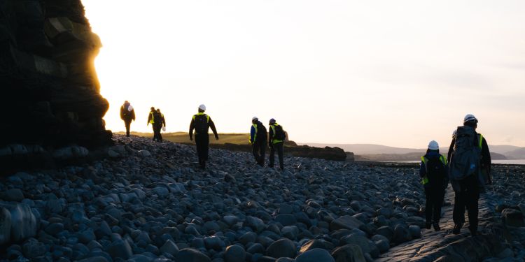 Researchers in hardhats and high vis jackets walk across a rocky terrain at sunset