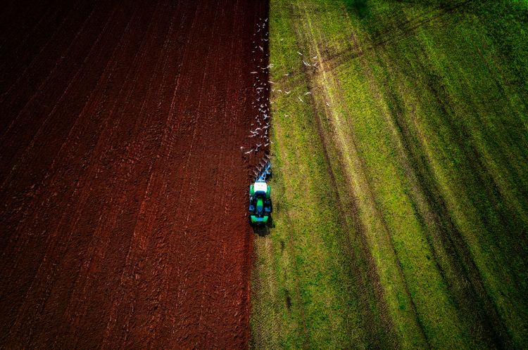An aerial view of a tracker ploughing a field. On the left is the brown soil, on the right is the grassy field.