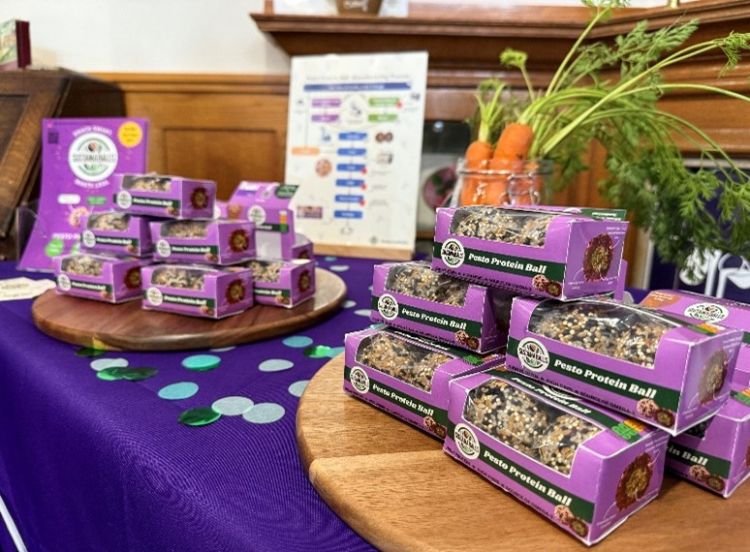 A display of SustainaBalls' product, pesto protein balls made from carrot tops and red grape skins. The products, small balls, are in small purple boxes. 