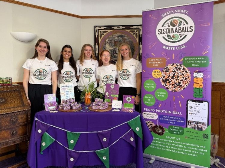 Team SustainaBalls behind their product table, wearing SustainaBalls t-shirts. The table and poster are purple and green. Their poster includes a picture of the product in its purple packaging and nutrition information.