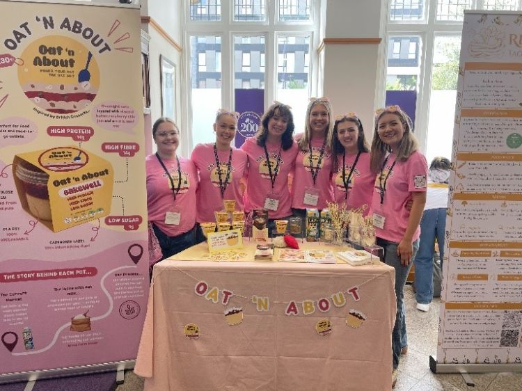 Team Oat 'N About with their product and poster. Team members are all wearing pink Oat 'N About t-shirts. Their poster includes a picture of the product in its pink and yellow packaging, nutrition information, and more details. 