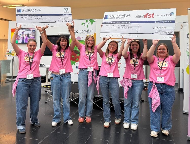 Winning team Oat 'N About holding up their prize money cheques, which are oversized and have Ecotrophelia UK's, Unilever's, IFST and Campbden BRI's logos on them as well. The team is smiling and wearing pink t-shirts and blue jeans. 