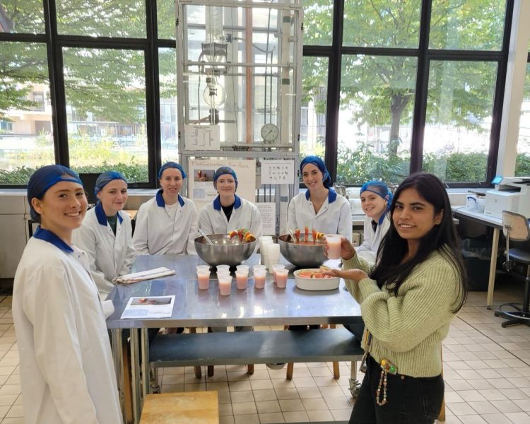 A group of people in lab coats and hair nets stood around a table with juice and cups on it.