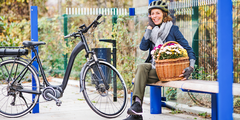 Someone sat on a bench next to an electric bike. They're wearing a bicycle helmet and holding a basket of flowers.