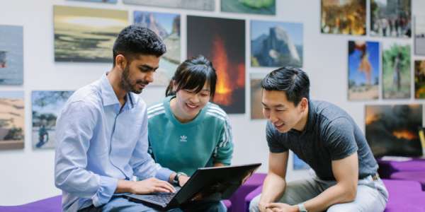 Three students sat around a laptop, behind them you can see posters of geological images on the wall.