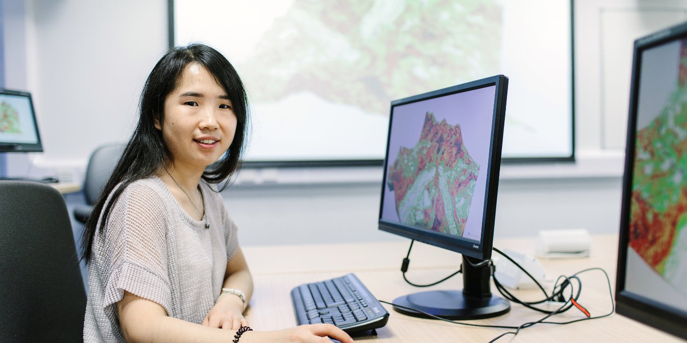 A student sat a desk in front of a desktop computer. On the screen is a geographical mapping image.