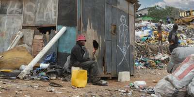 A Ghanaian person sitting on a stool. In the background we see a pile of used plastic and other rubbish.