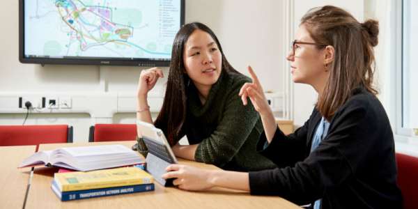 Two female-presenting students study together at a desk with a tablet, books and a large map on a screen in the background