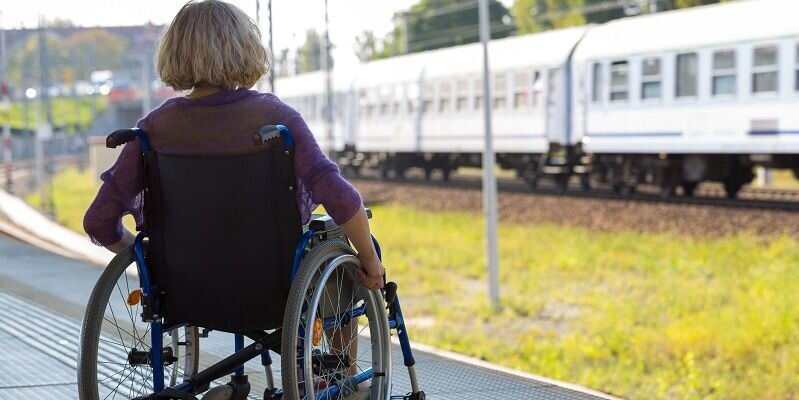 A woman in a wheelchair sat at a train station platform. There is a train passing in the distance.
